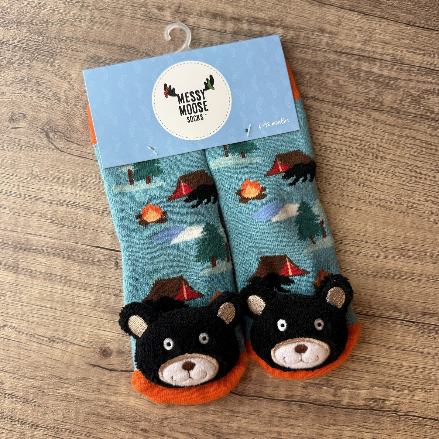 A pair of children's socks in a light blue woodland camping print, featuring 3D black bear faces on the toes and orange foot cuffs. The socks are packaged on a light blue hanger card labeled 'MESSY MOOSE SOCKS' and photographed flat on a wooden surface.