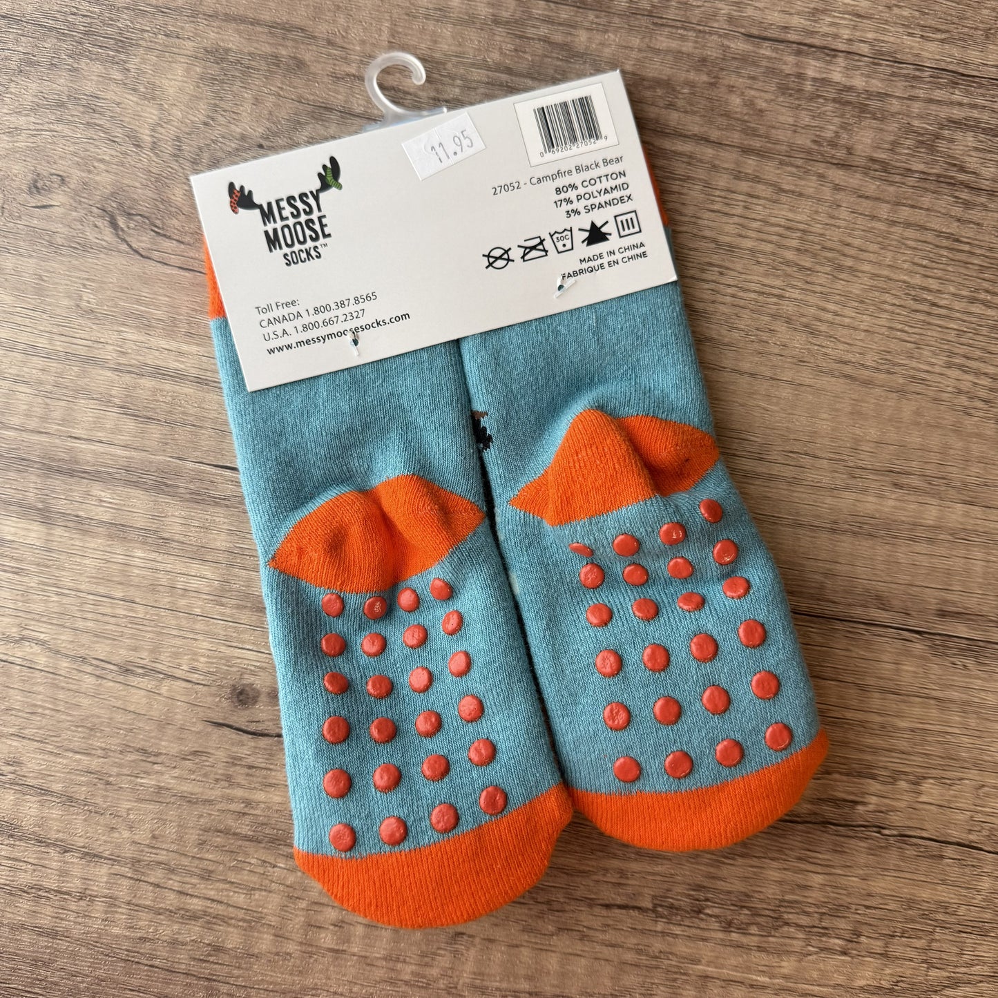 A pair of children's grip socks in teal blue and orange, packaged on a white cardboard hanger over a wooden floor. The socks feature textured orange grip dots and orange toe/heel sections.