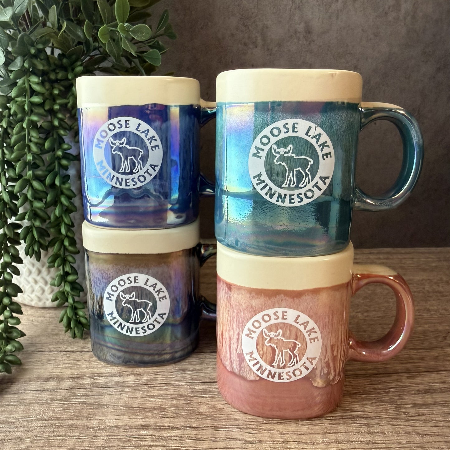 Four stacked ceramic mugs with Moose Lake Minnesota logos, displayed on a wooden surface with a green plant in the background. The mugs feature iridescent blue, teal, and purple glazes on the left stack, and a dusty pink, terracotta, and brown glaze on the right stack.