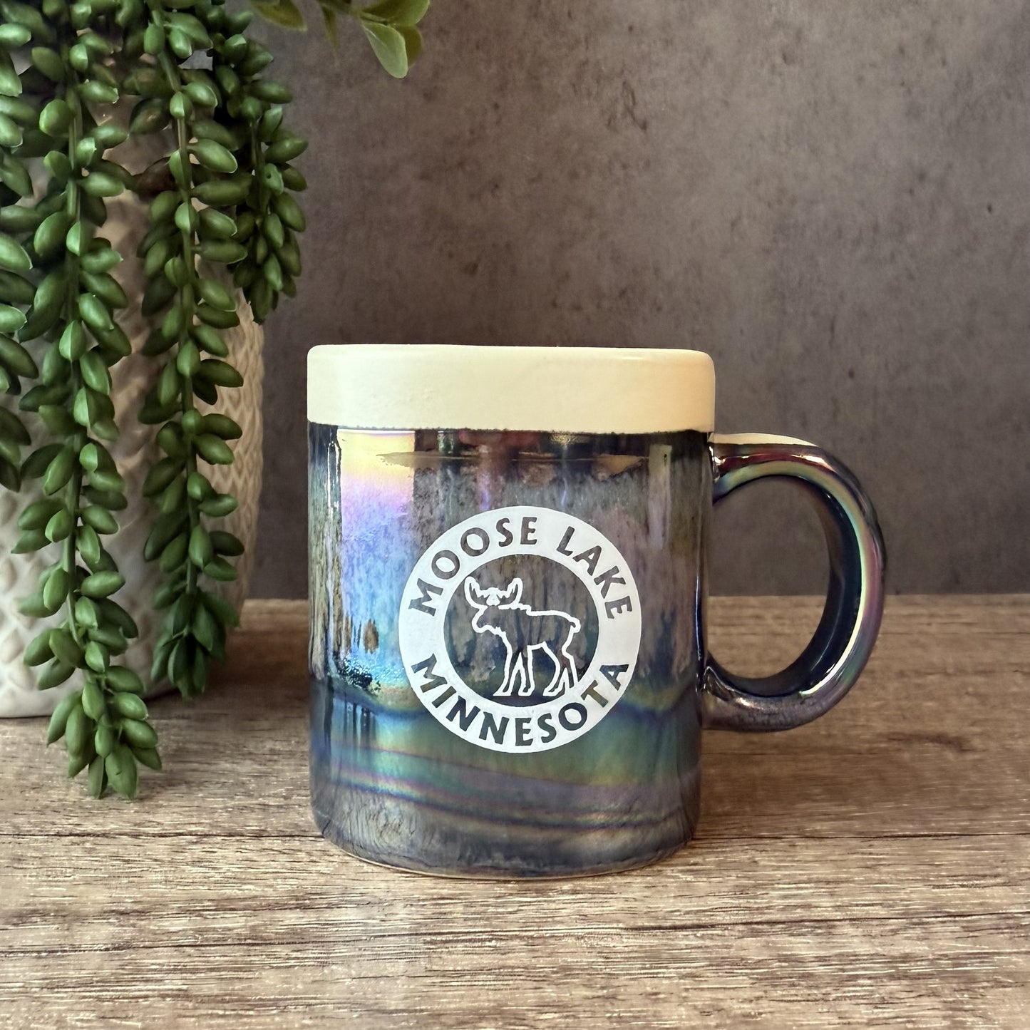 A ceramic mug with a light beige lid and a dark, iridescent body featuring a white circular 'Moose Lake Minnesota' logo. The mug sits on a wooden surface next to a potted green plant against a textured gray wall.