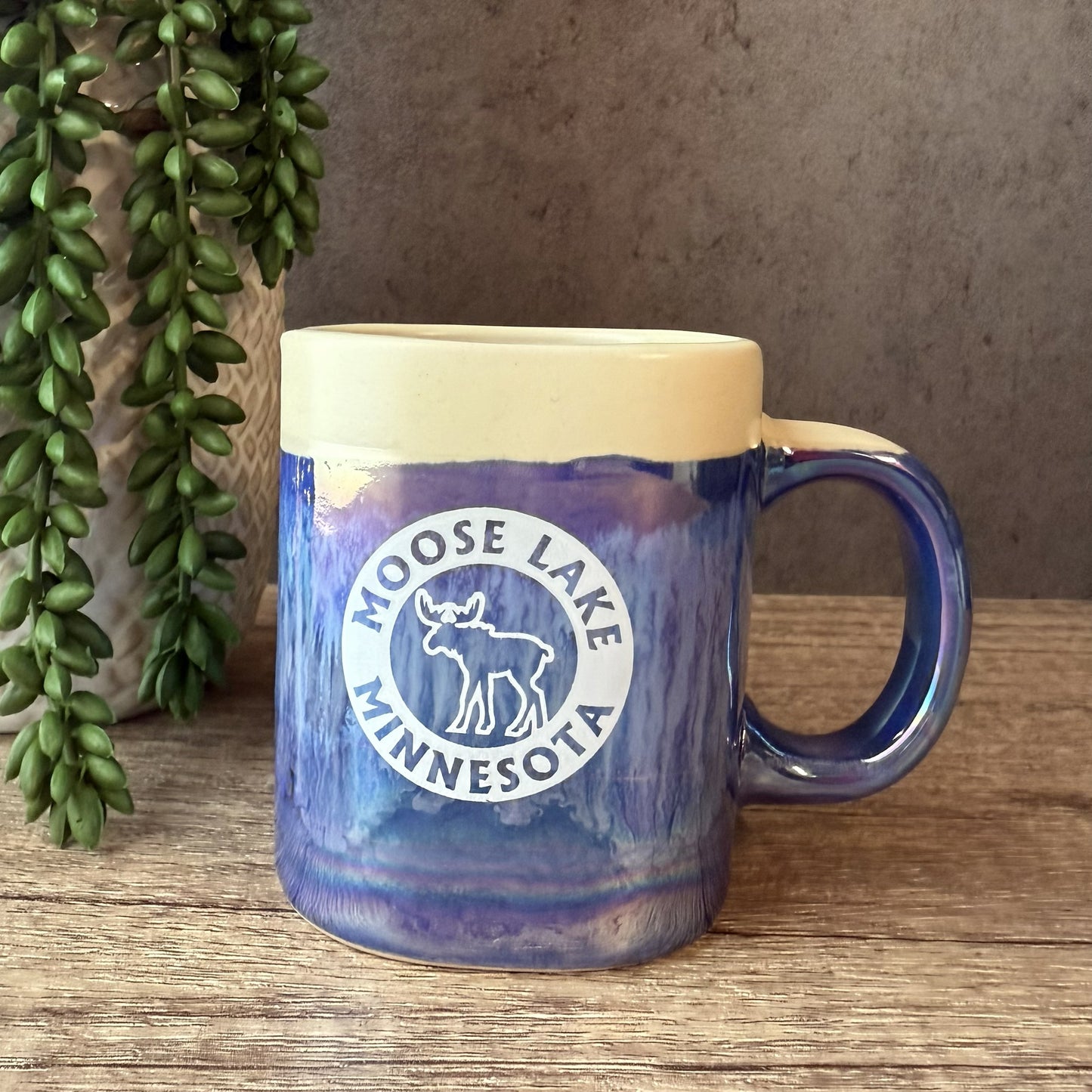 A ceramic mug with a blue and purple marbled pattern, a cream-colored interior, and a white circular 'Moose Lake Minnesota' logo on the front. The mug is placed on a wooden surface next to a hanging green plant.