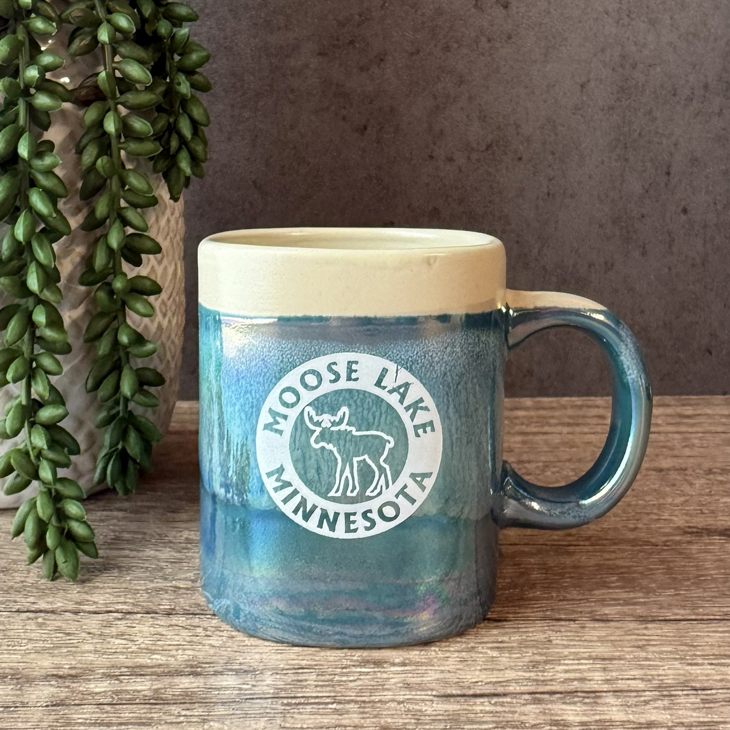 A ceramic mug with a blue and teal gradient body, a cream-colored rim, and a white circular 'Moose Lake Minnesota' logo. The mug is placed on a wooden surface next to a potted green plant against a textured gray wall.