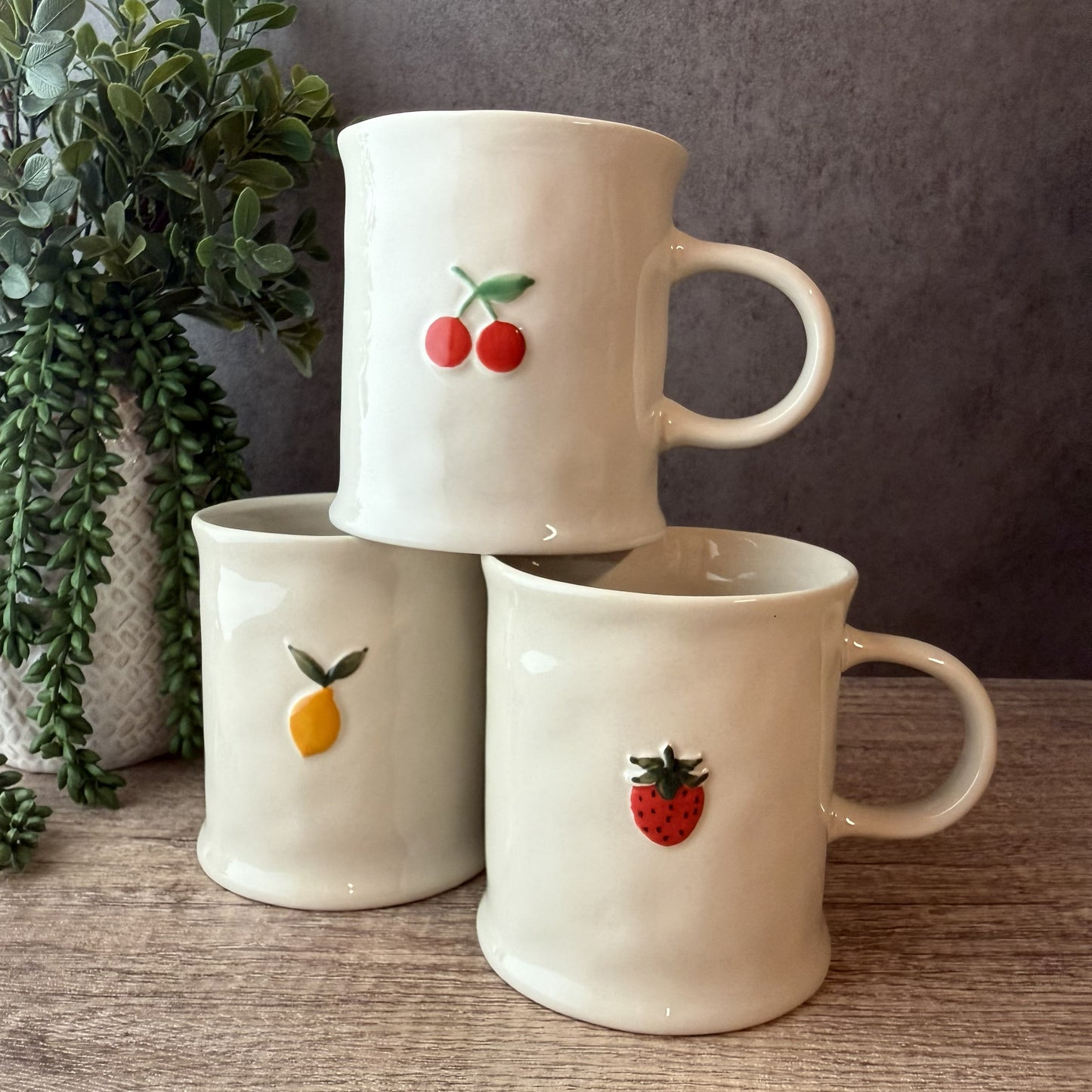 Three cream-colored ceramic mugs stacked together on a wooden surface, each decorated with a small fruit motif: cherries on the top mug, a yellow citrus fruit on the middle-left mug, and a strawberry on the middle-right mug. A potted trailing green plant is visible against a dark gray wall.