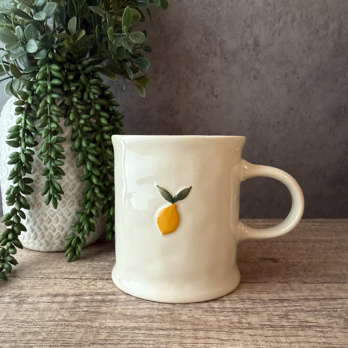 An off-white ceramic mug with a small 3D yellow lemon and green leaves design on the front, sitting on a wooden table next to a potted green plant against a gray wall.