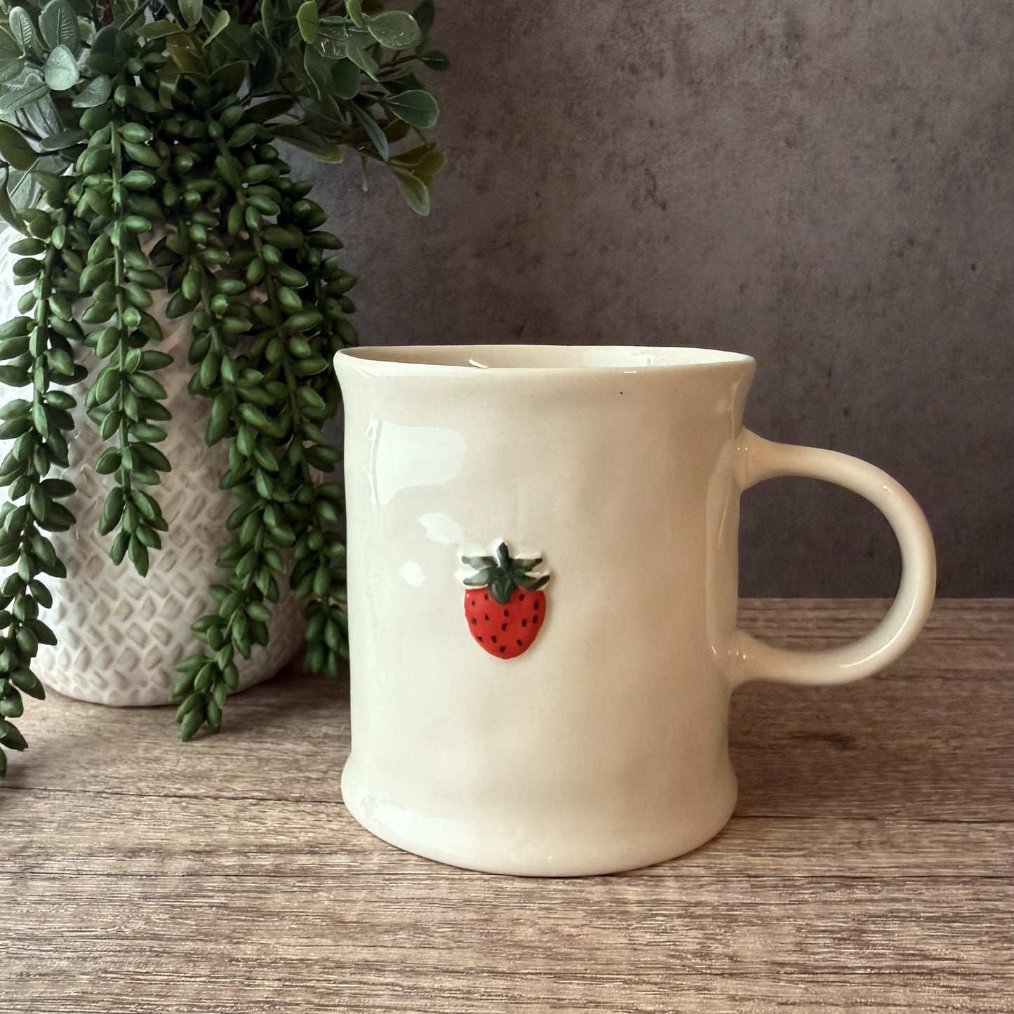 An off-white ceramic mug with a small red strawberry decoration on the front, sitting on a wooden surface next to a potted green plant against a gray wall.