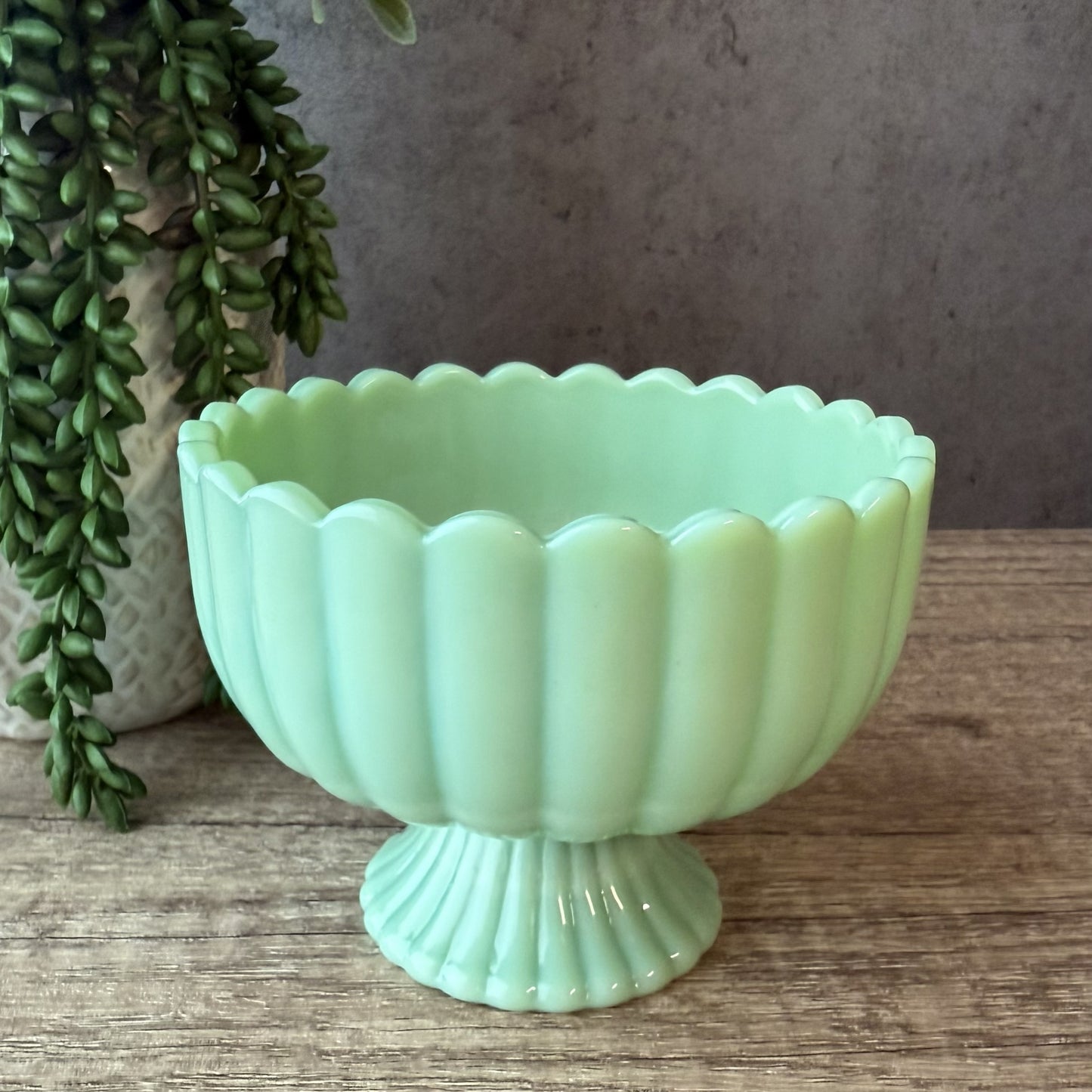 A light mint green glass pedestal bowl with a scalloped rim and ribbed base, displayed on a rustic wooden surface with a potted green plant in the background.