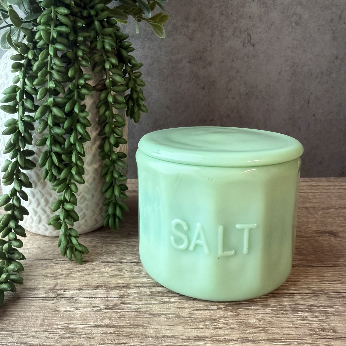 A mint green glass salt container with the word 'SALT' embossed on the front, sitting on a wooden surface next to a potted trailing green plant.
