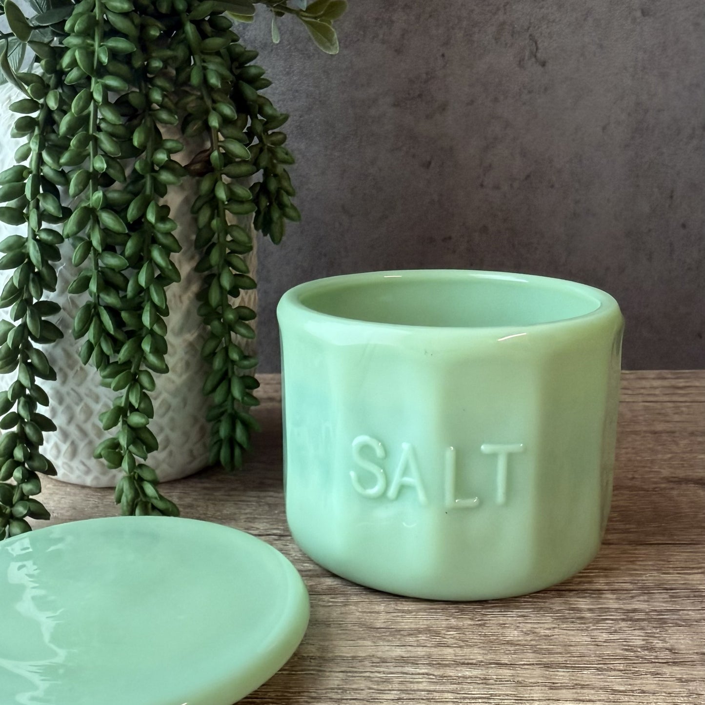 A mint green glass salt container with the word 'SALT' embossed on the front, shown next to its matching lid on a wooden surface, with a potted trailing green plant in the background.
