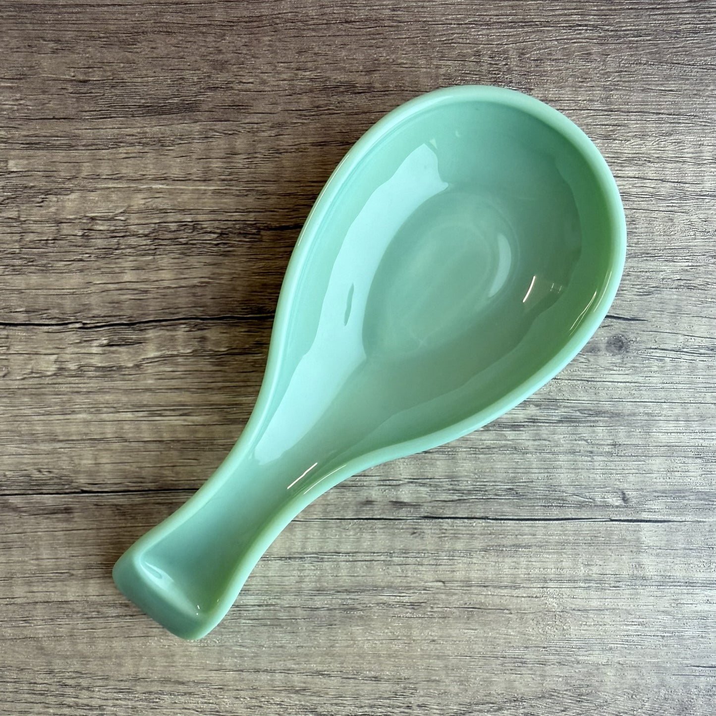 A glossy light green ceramic spoon rest with a teardrop shape, placed diagonally on a rustic wooden surface.