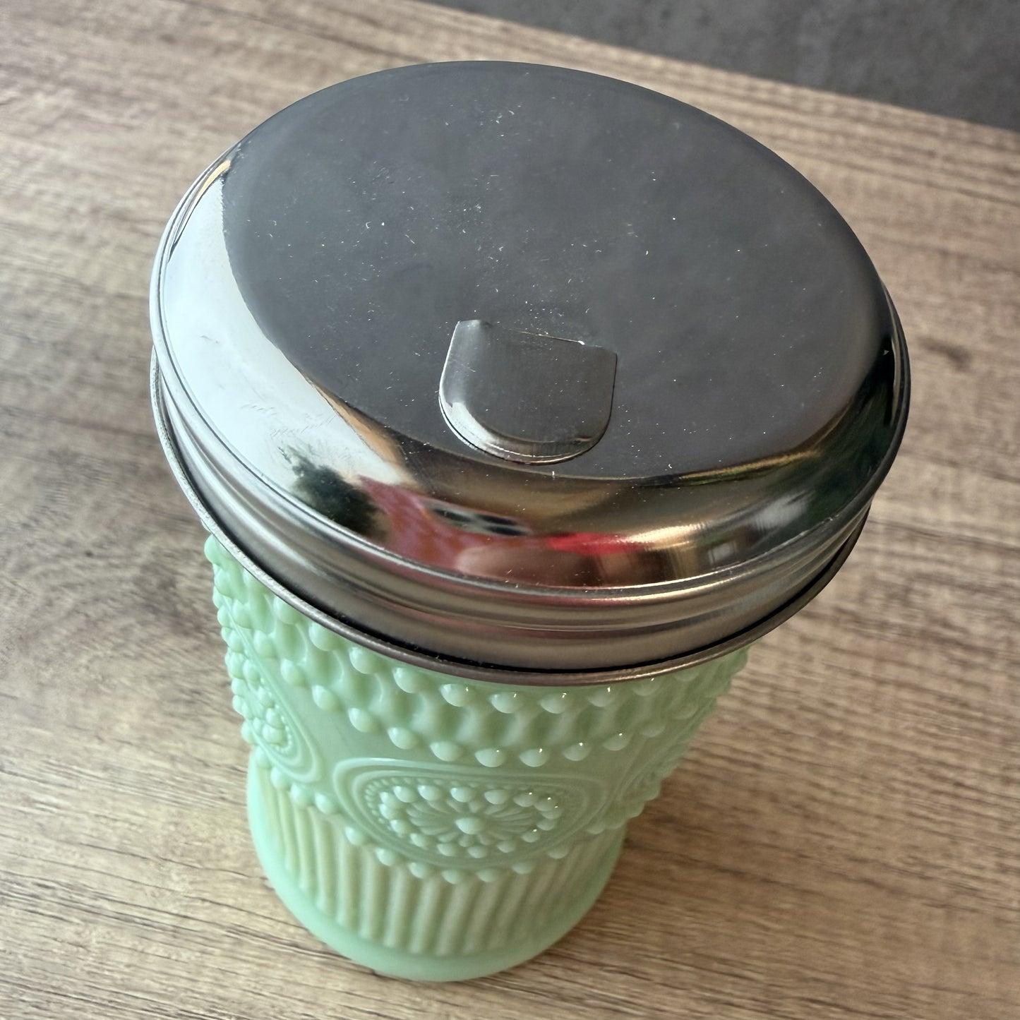A mint green reusable coffee cup with a textured honeycomb grip and a shiny stainless steel lid, resting on a wooden tabletop.