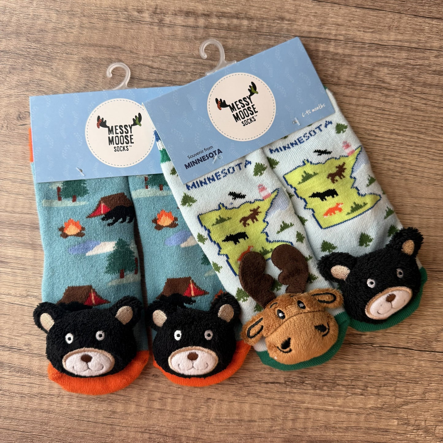 Three pairs of Minnesota-themed children's socks in their retail packaging, displayed on a wooden surface. The socks feature 3D animal footies: a black bear on orange, a moose with brown antlers, and a black bear on green.