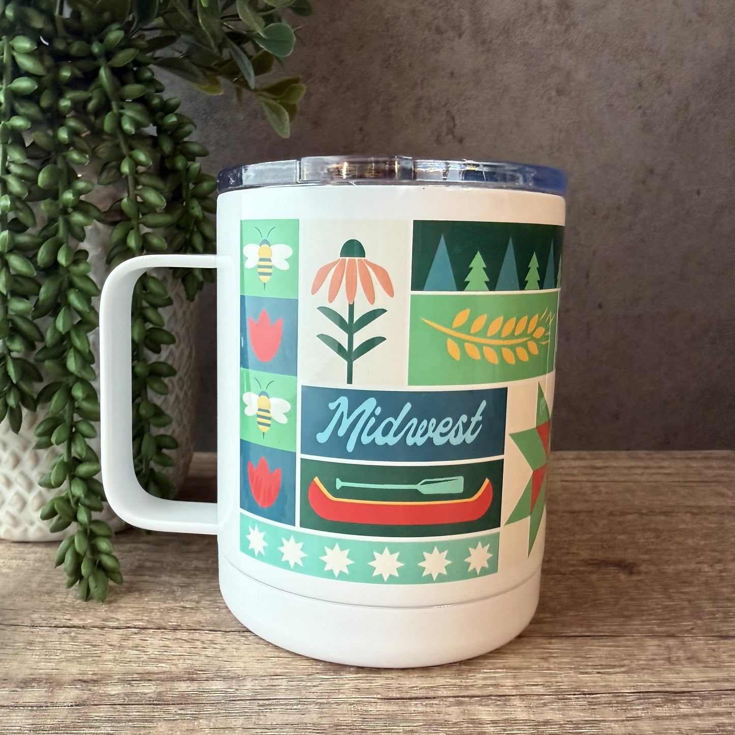 A white insulated handled tumbler with a clear lid sits on a wooden surface. The mug features a colorful Midwest-themed patchwork design with illustrations of bees, flowers, trees, wheat, a canoe, stars, and the word 'Midwest' in script.