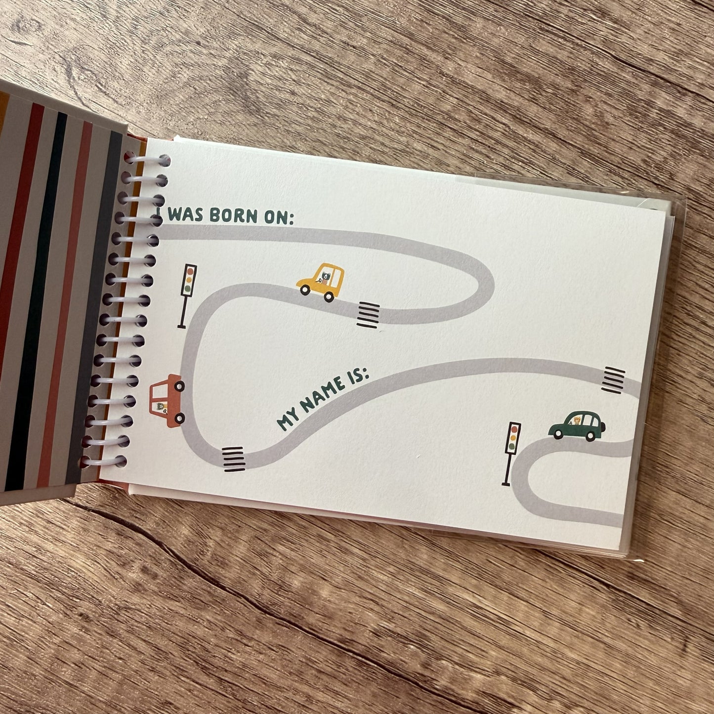 A spiral-bound notebook lying open on a wooden surface, showing a printed road-map style page with text reading 'WAS BORN ON:' and 'MY NAME IS:' plus small car illustrations.