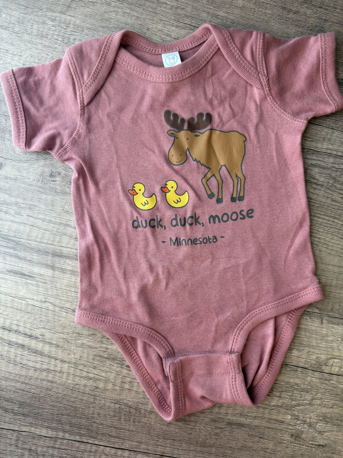 A dusty rose baby bodysuit with a graphic of a moose and two yellow ducks, accompanied by the text 'duck, duck, moose - Minnesota-', laid flat on a gray wood background.