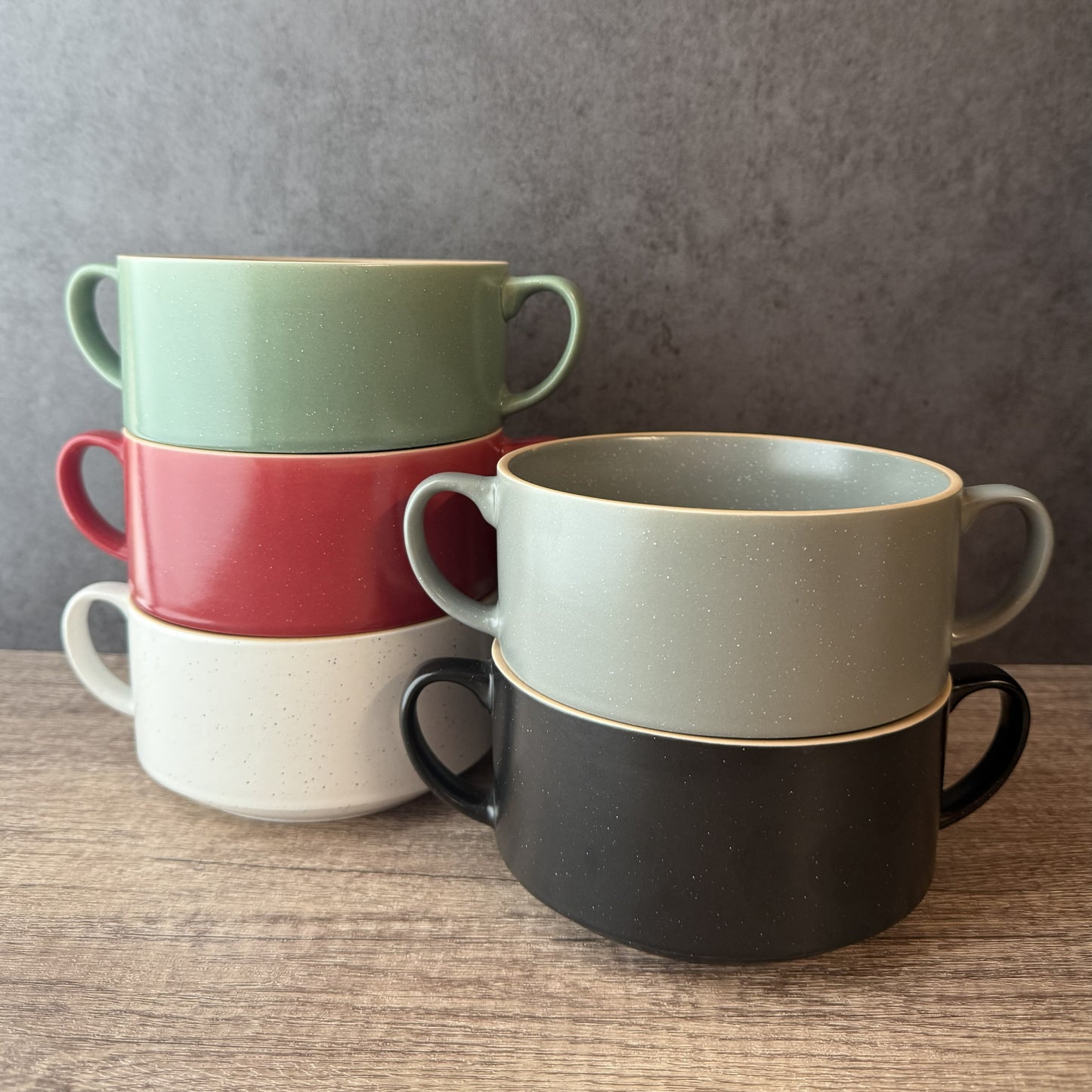 Five speckled ceramic bowls with handles, stacked in two groups of two, shown on a wooden surface against a gray textured background. The colors are sage green, coral red, off-white, and charcoal black.