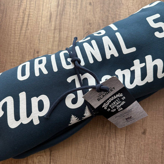 Rolled-up blanket with 'Original Up North' text on a wooden surface, featuring a tag.