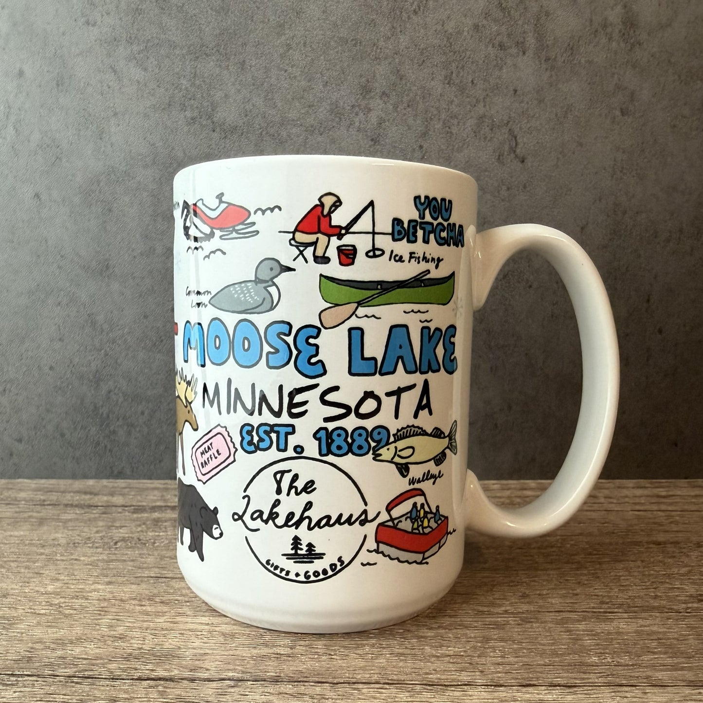 A white ceramic mug featuring hand-drawn illustrations and text related to Moose Lake, Minnesota, including 'YOU BETCHA Ice Fishing' and 'MOOSE LAKE MINNESOTA EST. 1869', placed on a wooden surface against a gray textured background.