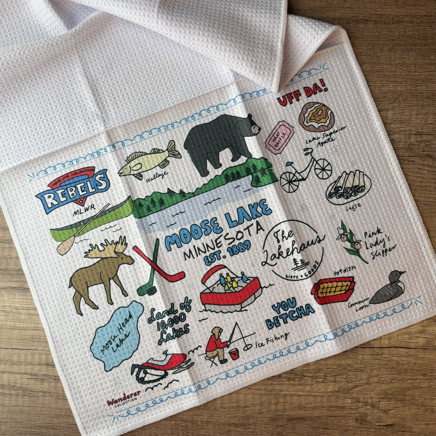 A white waffle-weave kitchen towel featuring colorful embroidered-style illustrations and Minnesota-themed phrases, laid flat on a wooden surface.